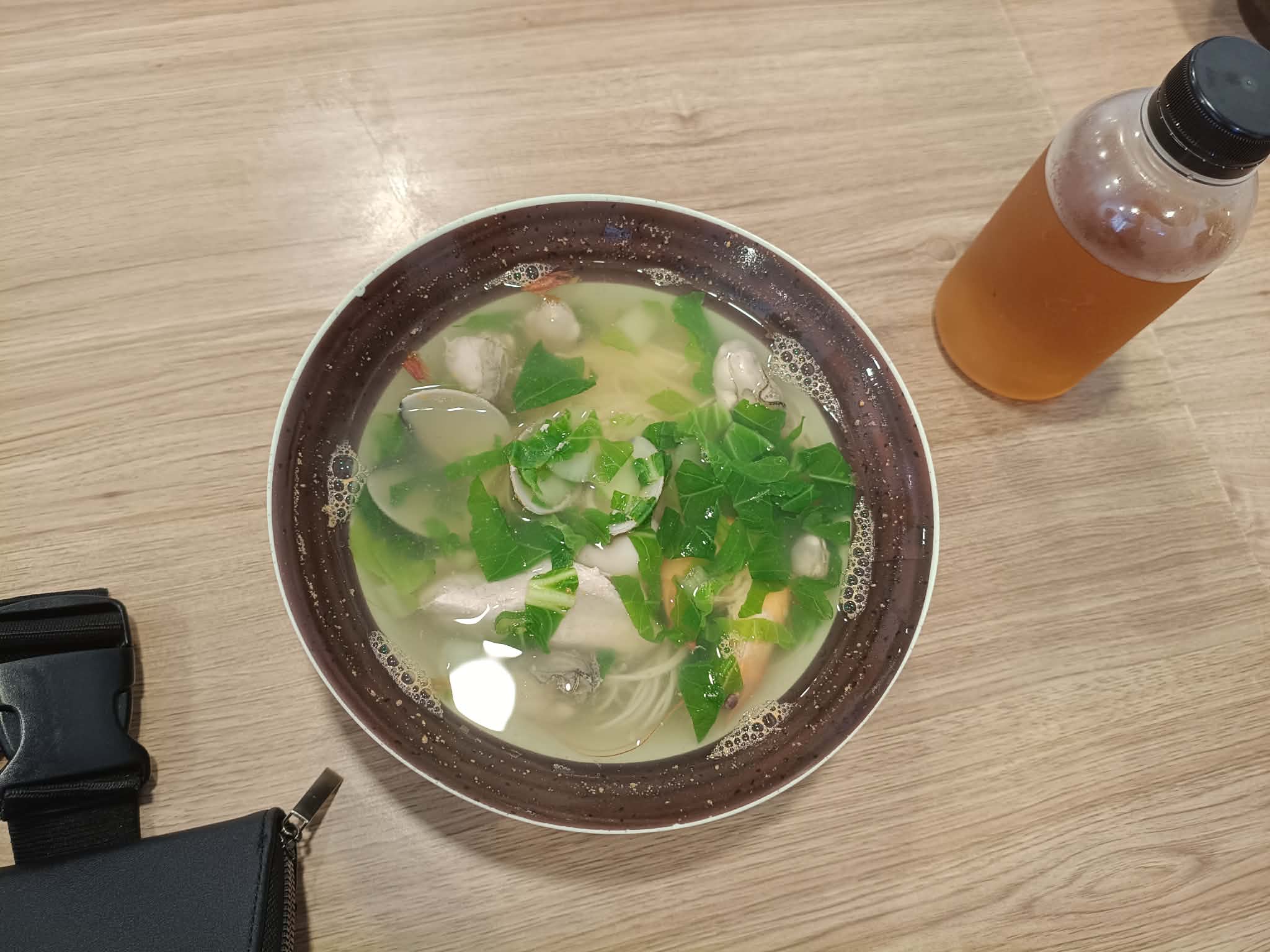 A bowl of clear seafood soup with clams, greens, and thin noodles, next to a bottle of iced tea